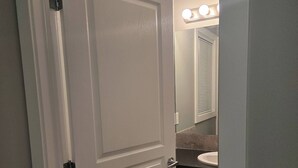 A closed white door with a light switch, a bathroom with a sink, mirror, and vanity lights. - Room-standard Bedroom With King Size bed (Lethbridge)
