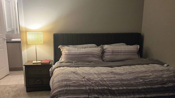 A bedroom with a bed, a bedside table with a lamp, and a nightstand. - Room-standard Bedroom With King Size bed (Lethbridge)