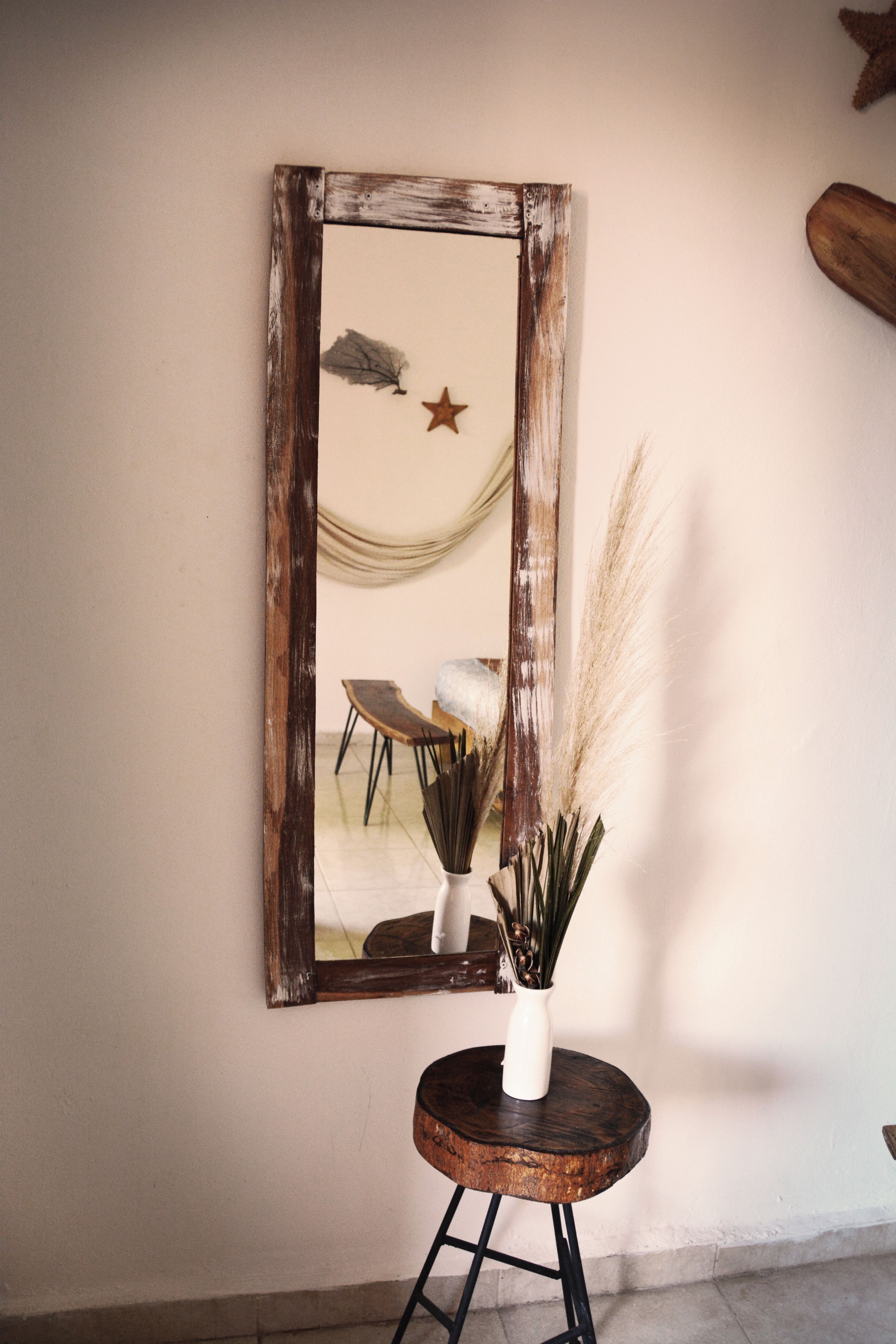 A rustic mirror with a distressed frame, a white vase with dried plants, and a wooden stool.