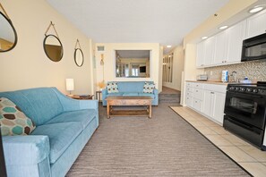 A living room with a blue sofa, a wooden coffee table, and a kitchen with white cabinets and a black oven. - 1BR Oceanfront on the Golden Mile (Myrtle Beach)