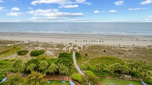 Une vue sur la plage, avec un sentier menant à un bâtiment, une plage de sable et quelques zones verdoyantes.