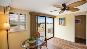A dining area with a glass table, wicker chairs, a ceiling fan, and a view of the outdoors through large windows. - Kona Alii 711 - Oceanfront Kona Penthouse w/ AC | Walk to Town (Kailua Kona)