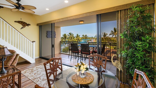 A glass-top dining table with wicker chairs, a ceiling fan, and a view of a pool and palm trees. - Kona Alii 711 - Oceanfront Kona Penthouse w/ AC | Walk to Town (Kailua Kona)