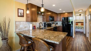 A kitchen with granite countertops, wooden cabinets, and stainless steel appliances. There are two wicker bar stools and a vase with branches. - Kona Alii 711 - Oceanfront Kona Penthouse w/ AC | Walk to Town (Kailua Kona)