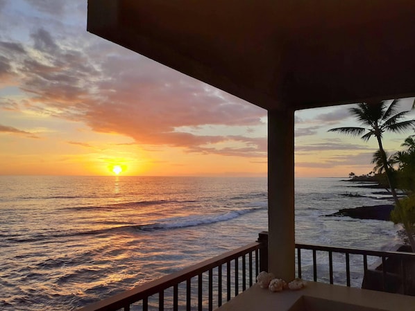A sunset view from a balcony with a railing, palm trees, and the ocean. - Banyan Tree 304 - Oceanfront Kona Condo w/ Views, Pool & Lanai (Kailua Kona)