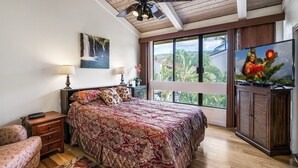 A bedroom with a bed, a nightstand, a lamp, a ceiling fan, a TV, and a window with a view of palm trees. - Surf & Racquet 3-301 Oceanfront 3BR Penthouse | Views, Lanai, AC (Kailua Kona)