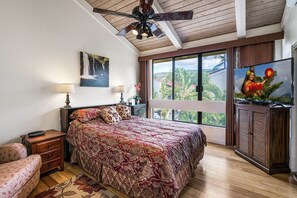 A bedroom with a bed, a nightstand, a lamp, a ceiling fan, a TV, and a window with a view of palm trees. - Surf & Racquet 3-301 Oceanfront 3BR Penthouse | Views, Lanai, AC (Kailua Kona)