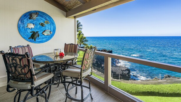 A dining area with a view of the ocean, a ceiling fan, and a decorative plate with sea creatures. - Surf & Racquet 3-301 Oceanfront 3BR Penthouse | Views, Lanai, AC (Kailua Kona)