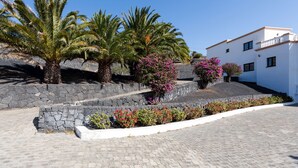 A paved area with palm trees and flowering plants, a stone wall, and a white building with balconies. - Bellavista private heated pool and sauna (Valles, Los)