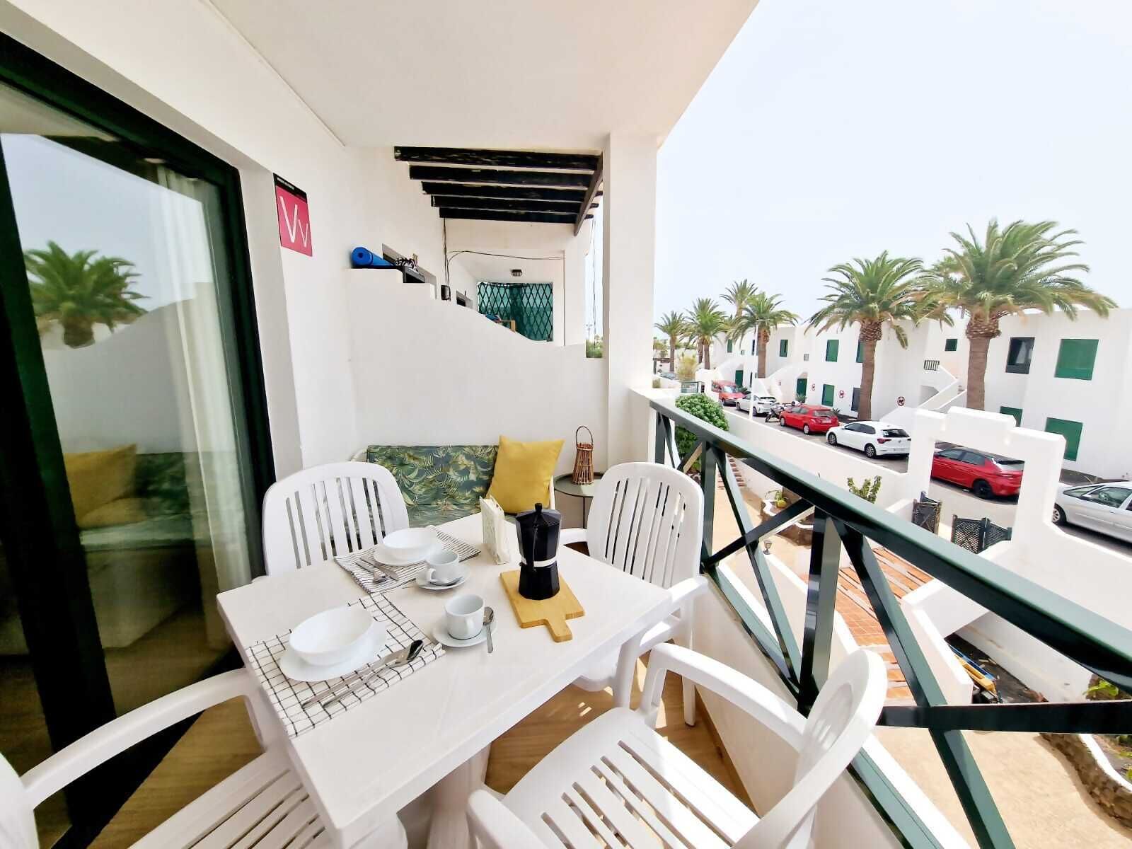 Ein Balkon mit Tisch für zwei Personen, weißen Stühlen, einem gelben Kissen und Blick auf eine Straße mit parkenden Autos und Palmen.