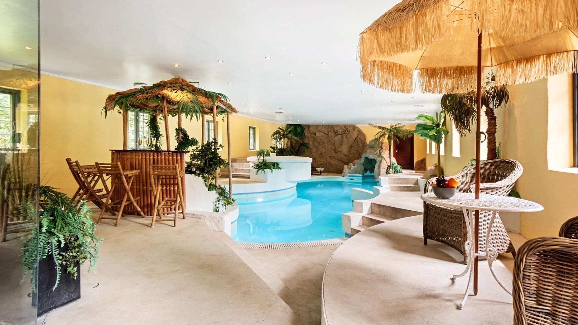 Un área de piscina con temática tropical, que incluye una sombrilla de paja, muebles de mimbre y abundante follaje.
