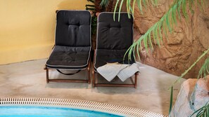 Two lounge chairs with cushions by a poolside, a white towel on one, and a rock wall with greenery in the background.