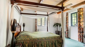 A four-poster canopy bed with a green patterned bedspread, a potted plant, and a window with floral curtains.