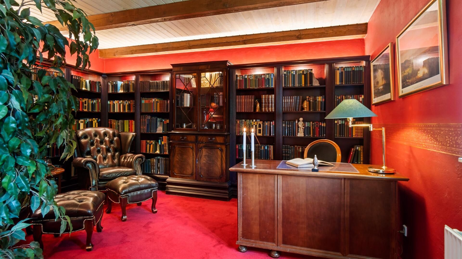 Una biblioteca con sillones de cuero, estanterías, un escritorio con lámpara y una alfombra roja.