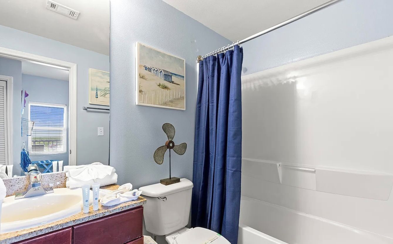 A bathroom with a white sink, a toilet, a blue shower curtain, and a fan.