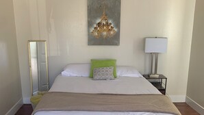 Un grand lit avec du linge de lit blanc, un oreiller vert à motifs, un plaid beige, un lustre, une table de chevet avec une lampe et un miroir au mur.
