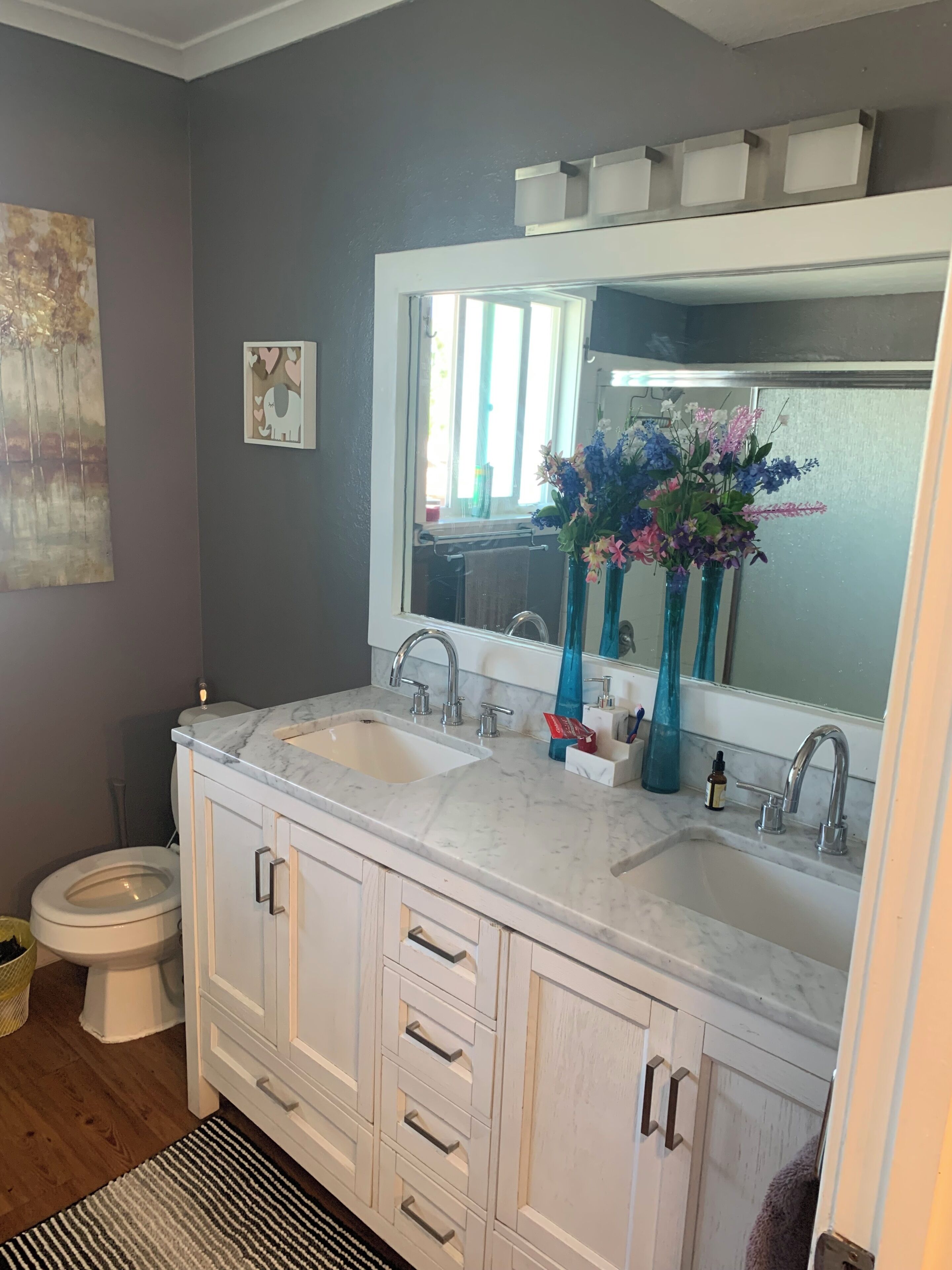 A bathroom with a double sink, a large mirror, a toilet, and a decorative painting on the wall.