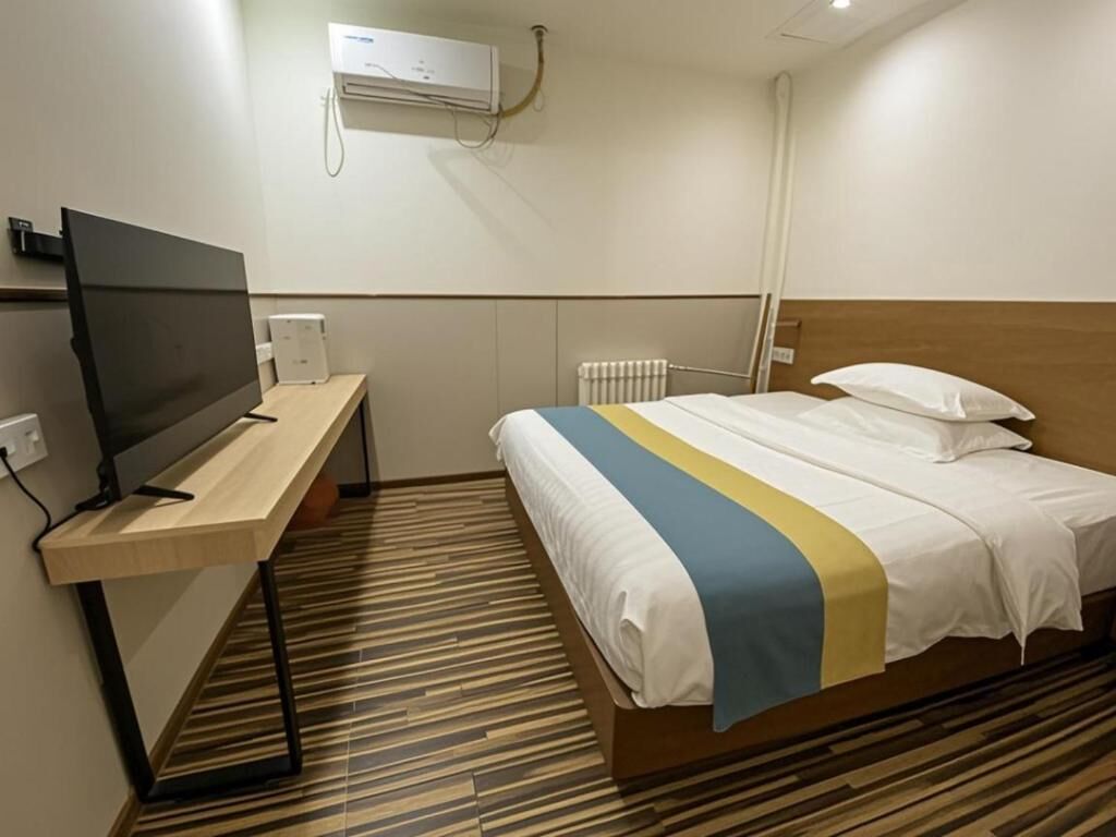 A hotel room with a bed, a desk with a flat-screen TV, a radiator, and a wall-mounted air conditioner.