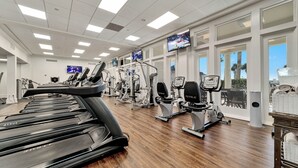 Un gimnasio con cintas de correr, elípticas y bicicletas estáticas, ventanas amplias y un televisor en la pared.