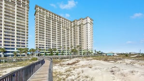 Un hotel frente a la playa con un paseo marítimo de madera que conduce a la arena, edificios altos y palmeras.