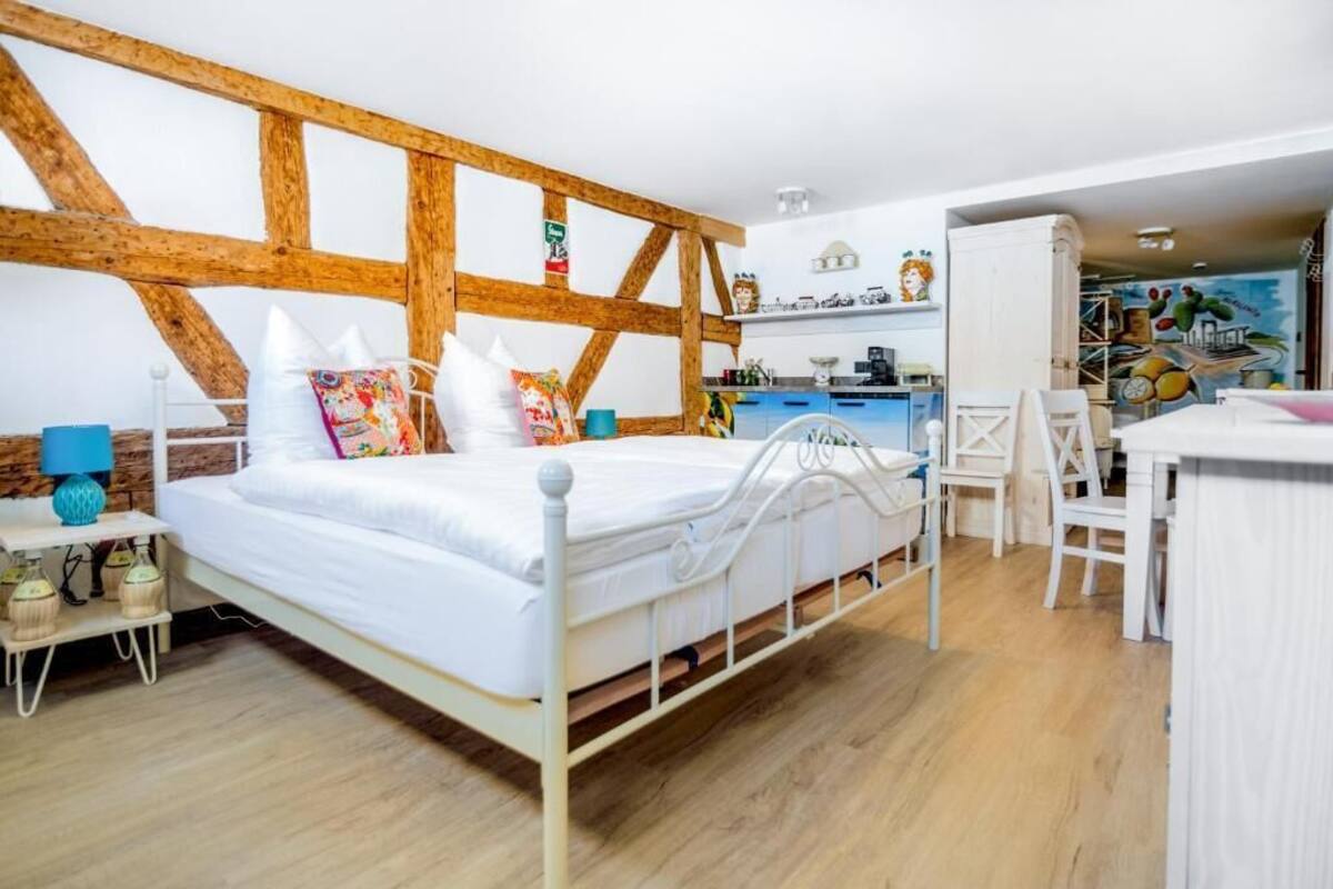 A bedroom with a bed, bedside table, lamp, and a view of the kitchen area.