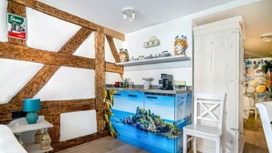 A room with exposed wooden beams, a kitchen area with a blue island, a white chair, and a shelf with decorative items. - H M Boarding Houses (Lauf an der Pegnitz)
