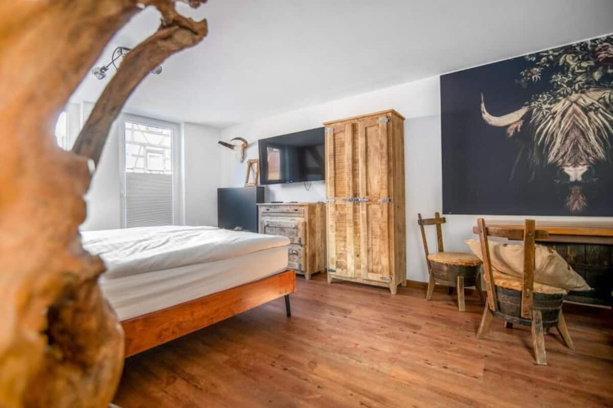 A bedroom with a wooden bed, a desk with a chair, a television, and a large painting on the wall.