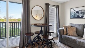 A balcony with a table and stools, a grey sofa with patterned cushions, a round mirror, and a bull painting on the wall. - Riverfront Rhythm by Avantstay Shared Pool + Balcony + River Views Walk to Broadway + Germantown (Nashville)