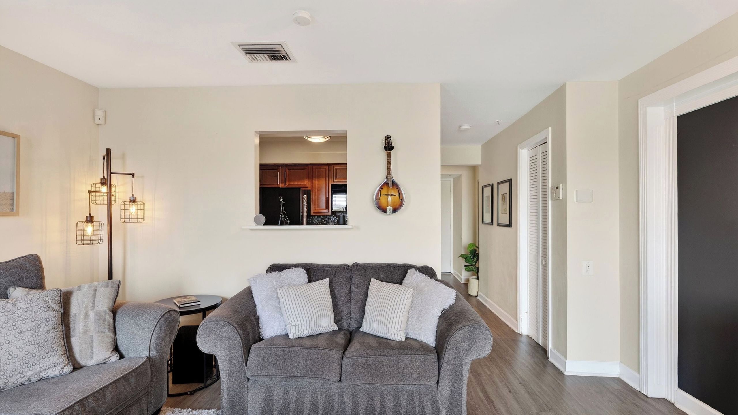 Una sala de estar con un sofá gris, una mesita y una lámpara. Se ve una cocina a través de un vano abierto.