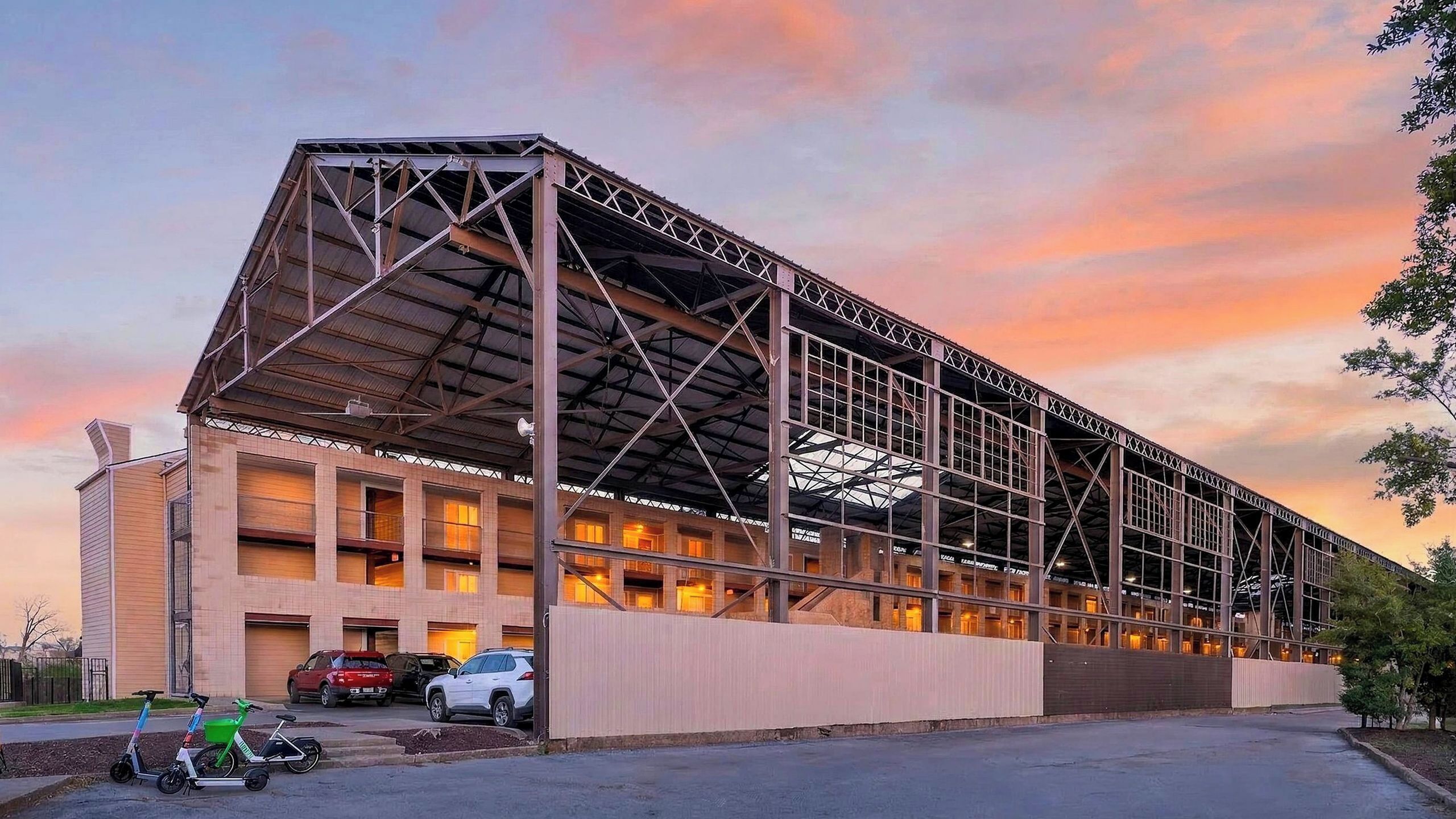 A large industrial-style building with a metal framework, multiple windows, and a parking area with cars and scooters.