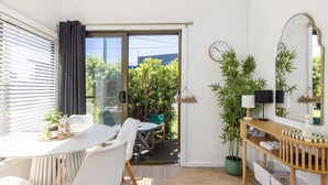 Une pièce dotée d’une porte-fenêtre coulissante donnant sur un jardin, d’une table blanche avec des chaises, d’une console en bois avec des lampes et d’un miroir au mur.