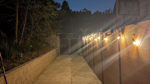 A paved pathway leading to a basketball court with a low wall and string lights.