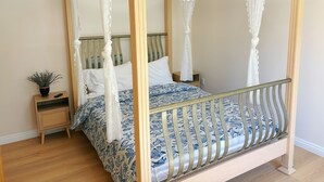 A four-poster canopy bed with a wooden frame and white drapes, a bedside table with a plant, and a patterned bedspread.