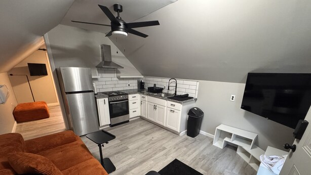 A compact kitchen with a stove, oven, and sink, a ceiling fan, a TV mounted on the wall, and a sofa with a remote on a small table. - A Little Country w/ A Little Beach (BOLIVIA)