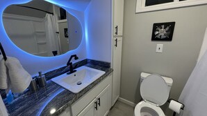 A bathroom with a white sink, a mirror with a blue light, a toilet, and a fan on the wall. - A Little Country w/ A Little Beach (BOLIVIA)