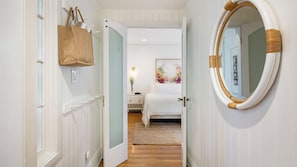 A hallway leading to a bedroom with a bed, a nightstand, a lamp, a mirror, and a door with a handle.