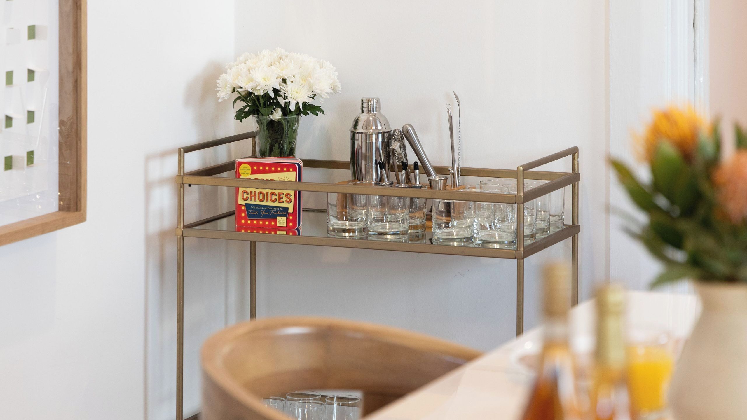 A bar cart with a boxed set of cocktail recipes, a vase of flowers, and various bar tools.