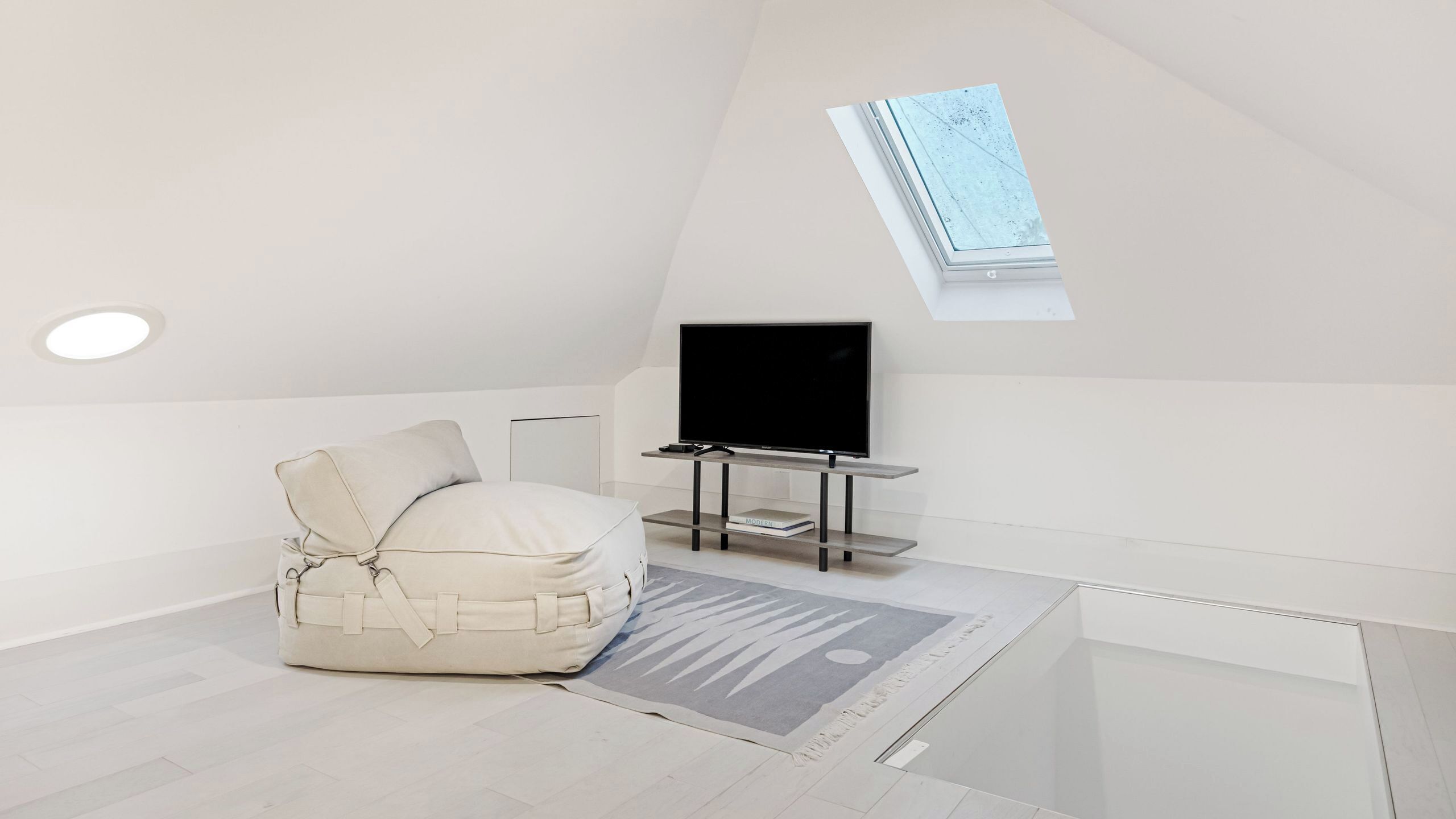 Une pièce moderne et minimaliste avec un plafond en pente, une grande fenêtre, un téléviseur à écran plat posé sur un meuble, un coin salon confortable et un tapis à motifs.