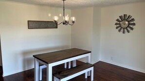 Un comedor con una mesa y banco de madera, una lámpara de araña, un reloj de pared y un cartel enmarcado.