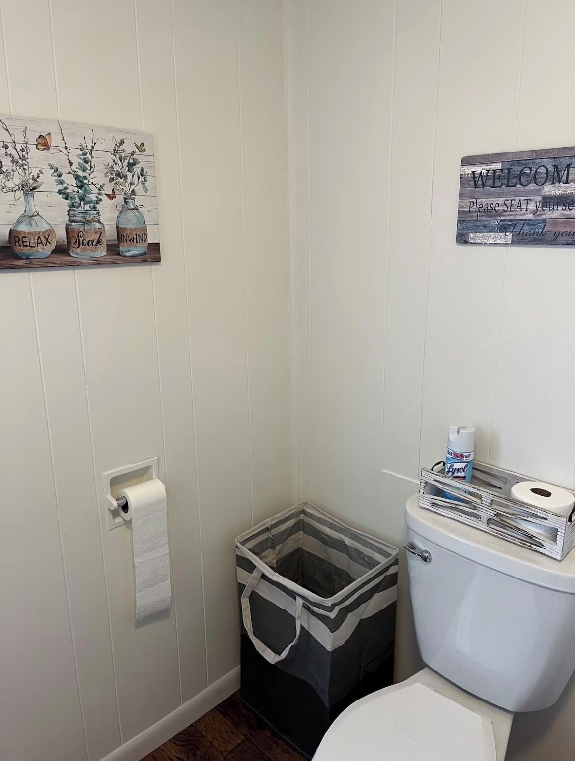 A bathroom with a toilet, a roll of toilet paper, a trash bin, and a welcome sign.