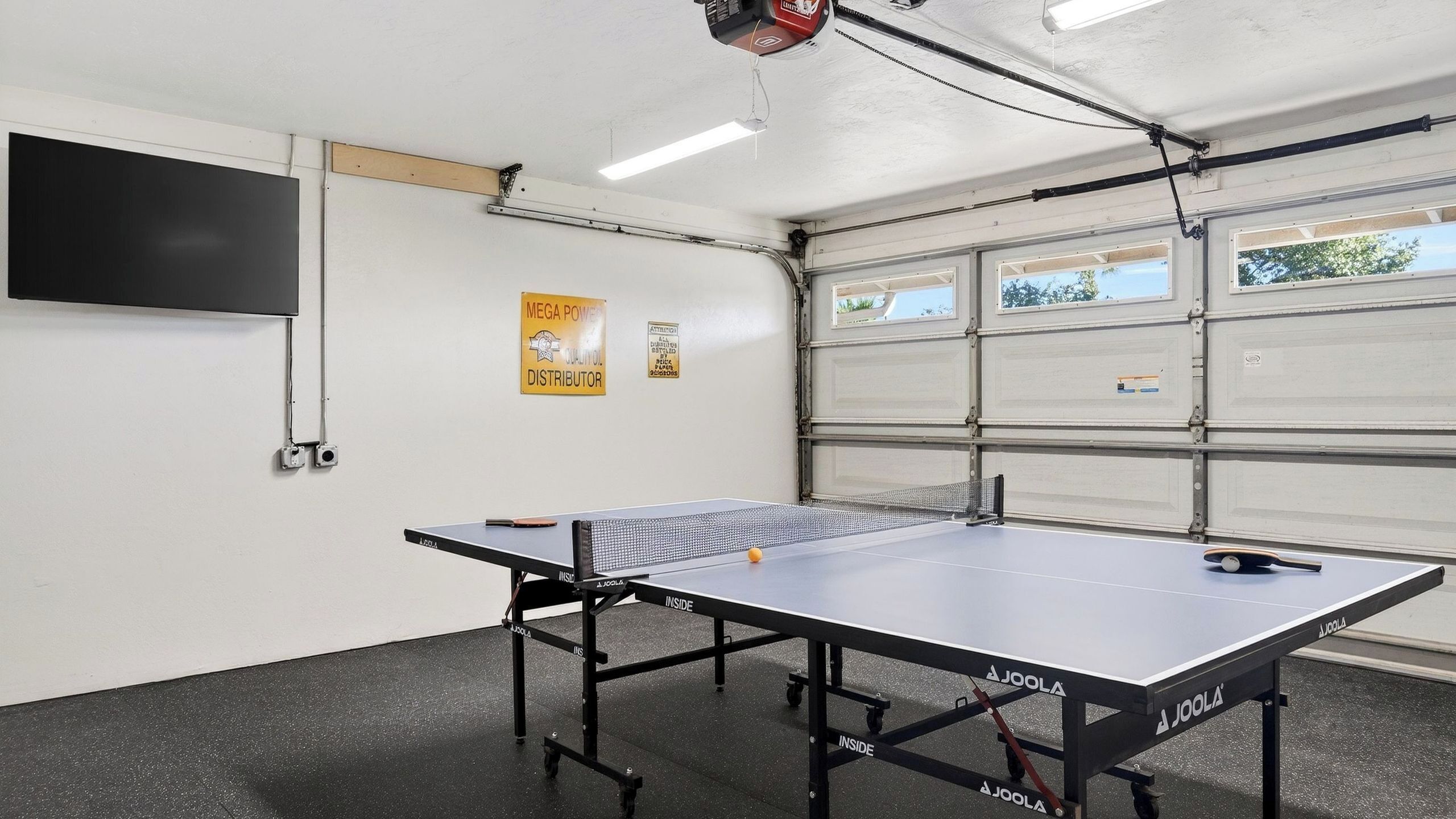 Une table de ping-pong dans un garage, avec une télévision et une affiche accrochée au mur.