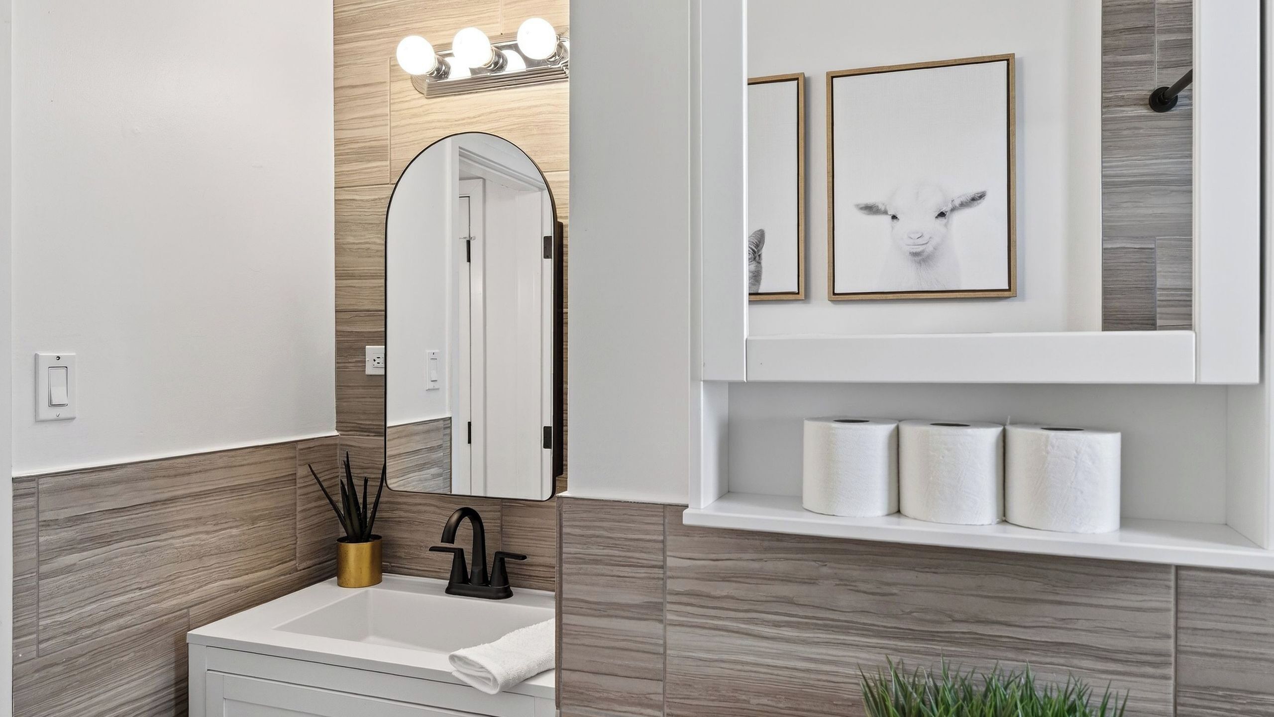 Un baño moderno con un lavamanos blanco, grifo negro y un espejo con luces arriba. Hay cuadros enmarcados en la pared y una repisa con papel higiénico.