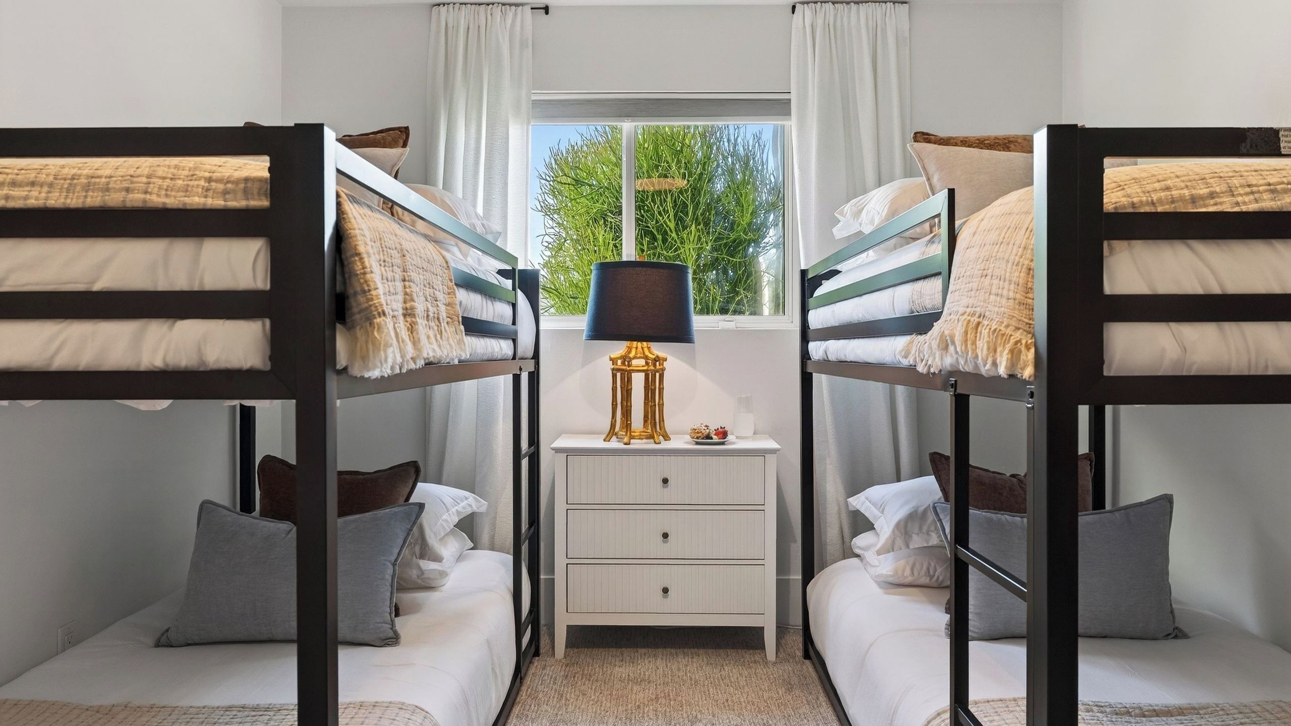 A bunk bed room with a desk, drawers, a lamp, and a window with a view of greenery.
