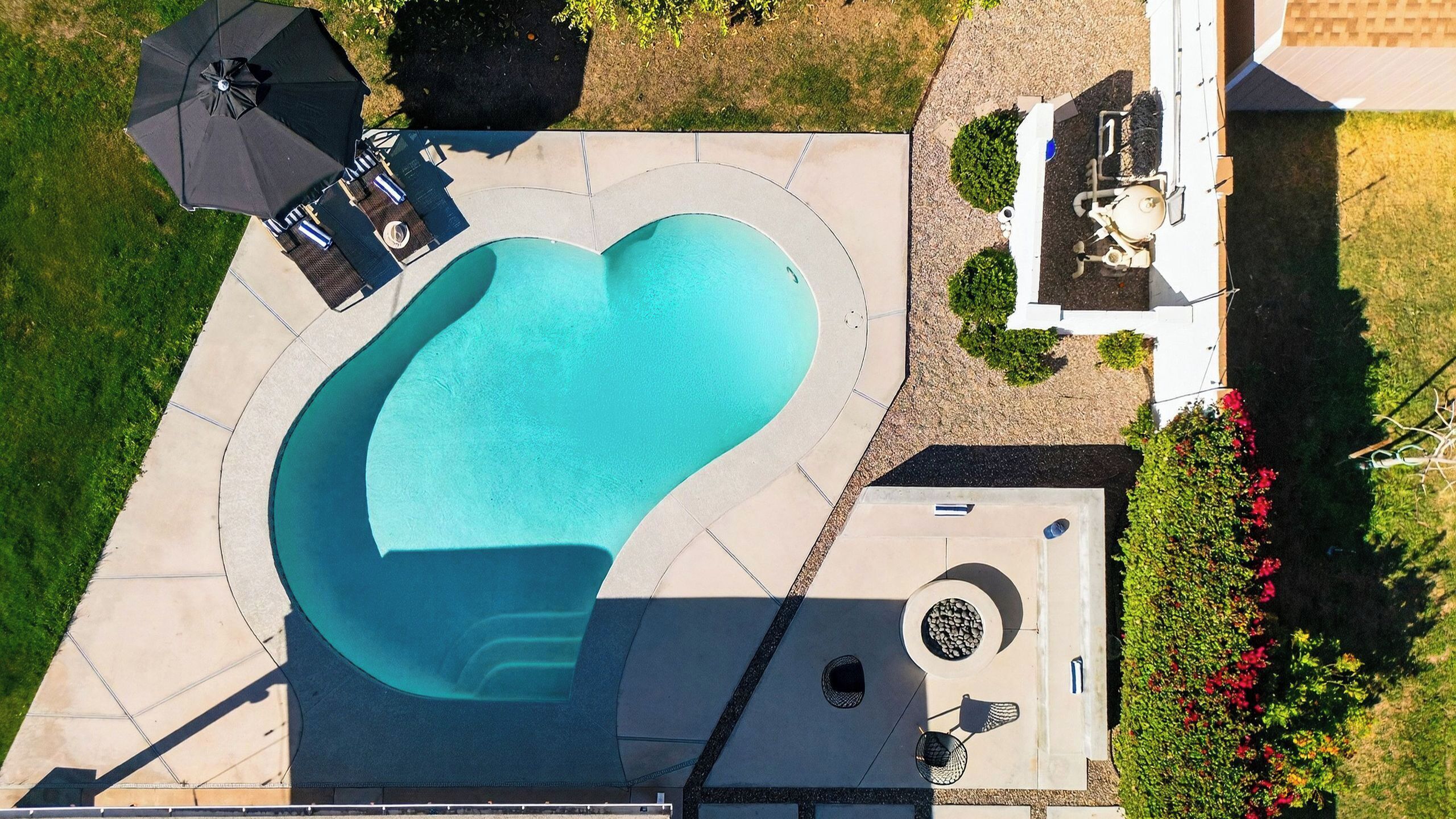 Vue aérienne d’une piscine en forme de cœur, entourée d’une terrasse aménagée avec des chaises longues et des parasols.