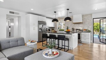 Una cocina moderna con una isla central, taburetes de bar y una zona de comedor con un sofá gris y una pequeña mesita auxiliar.