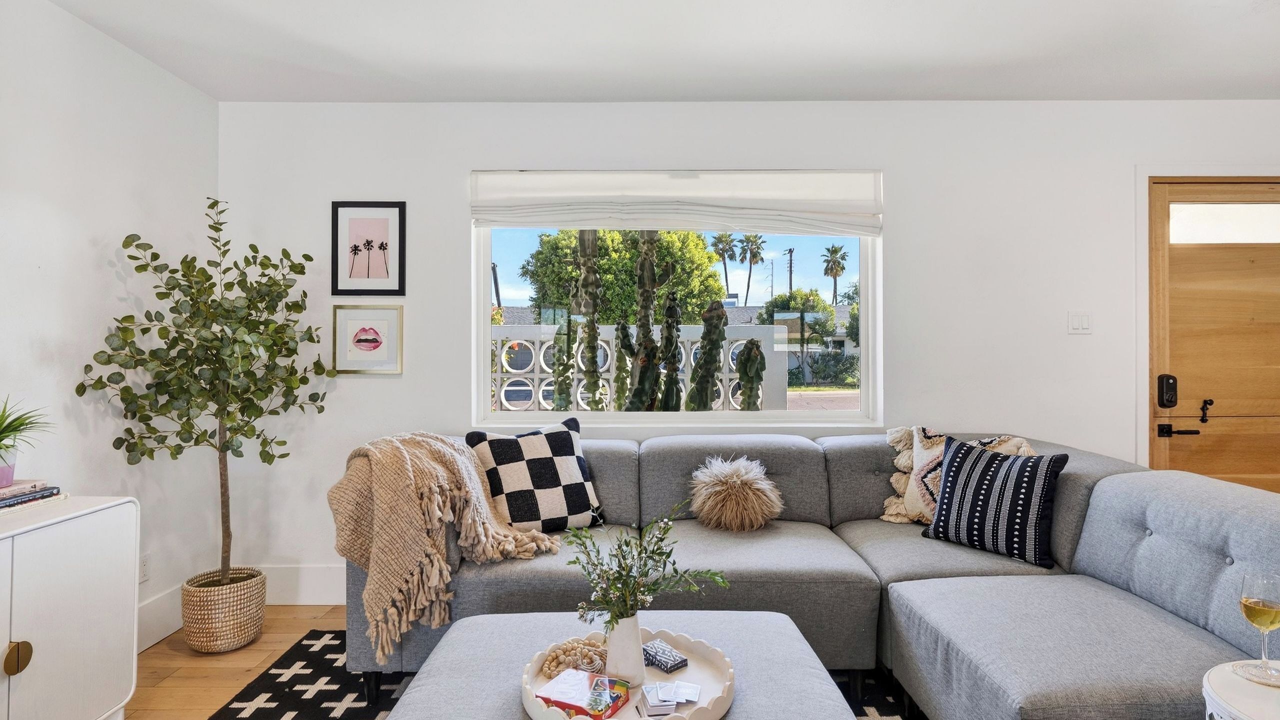 A modern living room with a grey sectional sofa, a coffee table with decorative items, a large window with a view of trees and a fence, and a wooden door.