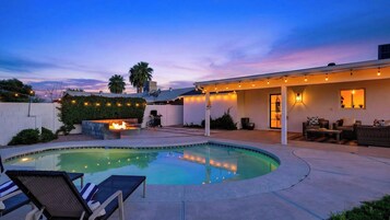 Un jardín trasero con piscina, una fogata, mobiliario exterior e iluminación colgante.