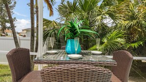 Ein Tisch mit Glasplatte, Korbstühle, eine blaue Vase und zwei Weingläser auf einem Balkon mit Palmen und einem weißen Zaun im Hintergrund.
