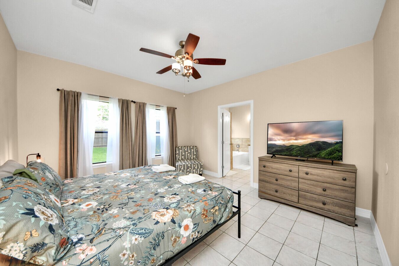 A bedroom with a bed, a ceiling fan, a TV, a dresser, and a bathroom visible through an open door.