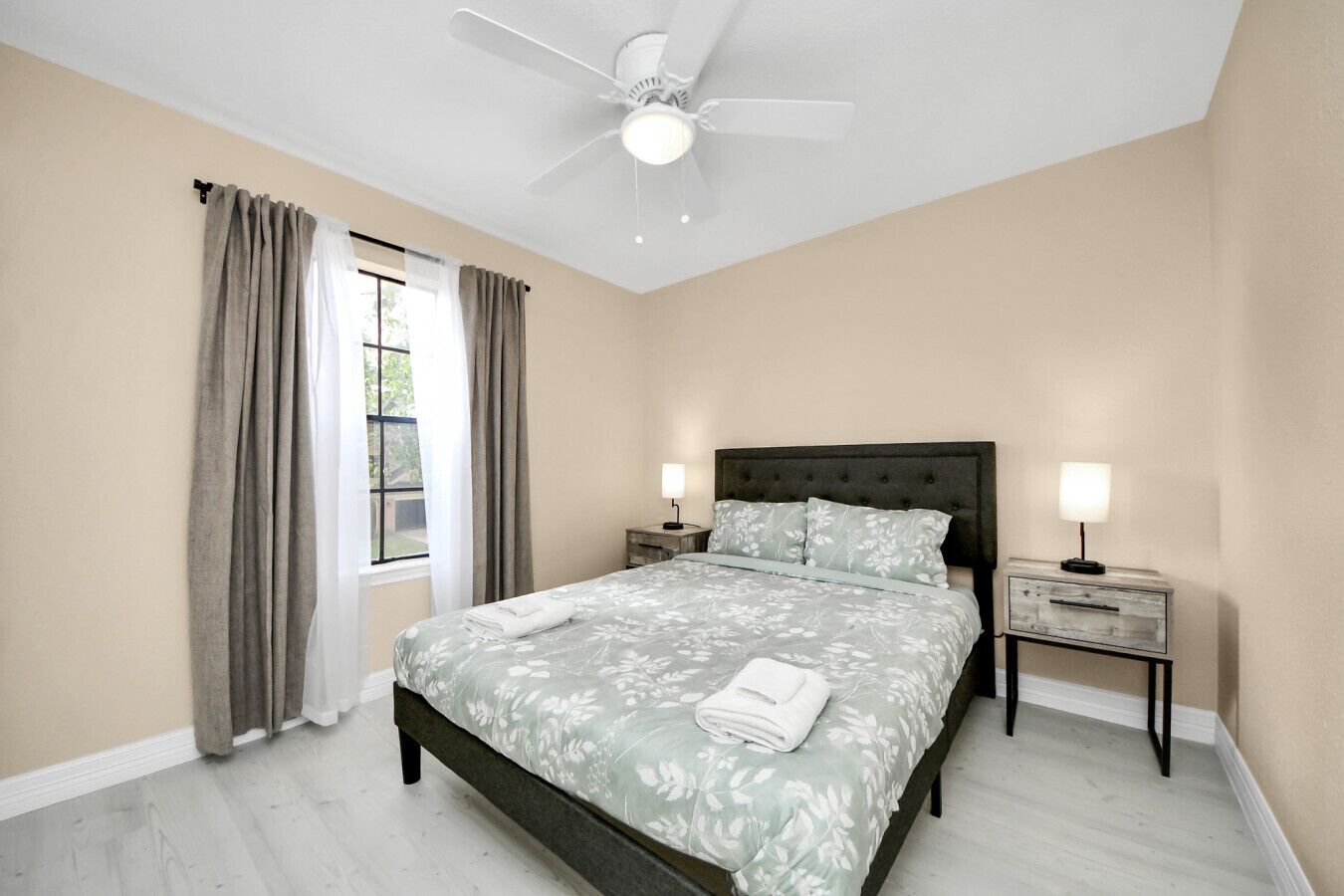 A bedroom with a bed, a ceiling fan, a nightstand with a lamp, and a window with curtains.
